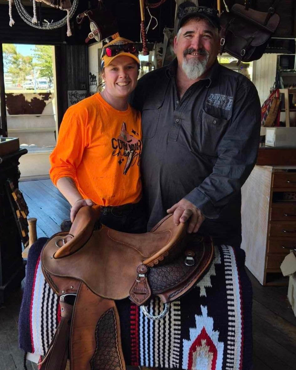 Two people holding a saddle in a rustic indoor setting